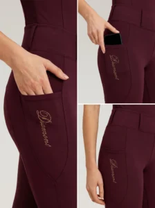 Close-up details of burgundy riding leggings: side phone pocket holding a smartphone and Diamont script