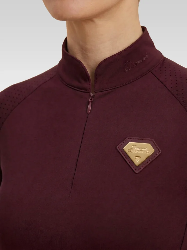 Close-up of the burgundy Prestige long sleeve base layer: stand-up collar, quarter-zip, perforated shoulder panel and gold metal badge in a burgundy leather frame