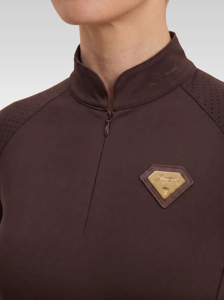 Close-up of the chocolate Prestige long sleeve base layer: stand-up collar, quarter-zip, perforated shoulder panel and gold metal badge in a brown leather frame
